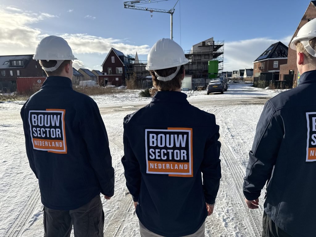 Drie vakmensen van Bouwsector Nederland met witte helm kijken uit over een nieuwbouwwijk met een bouwkraan